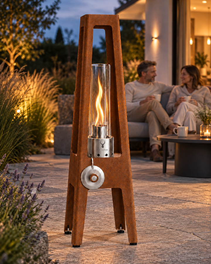 TORRE Corten bioethanol lamp with metal legs in the foreground of an outdoor seating area with people farther behind.